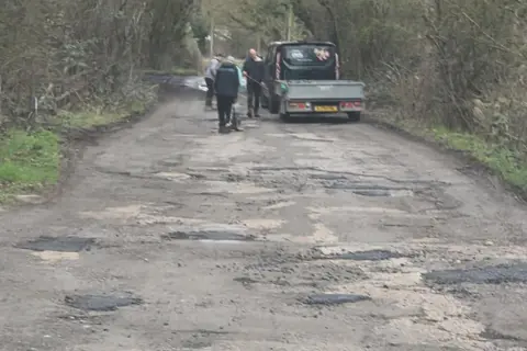 Michelle Thexton A photo of Pete Thexton and two other men working from a van as they fill potholes along Aldercar Lane. In the foreground darker patches of freshly placed tarmac are visible.