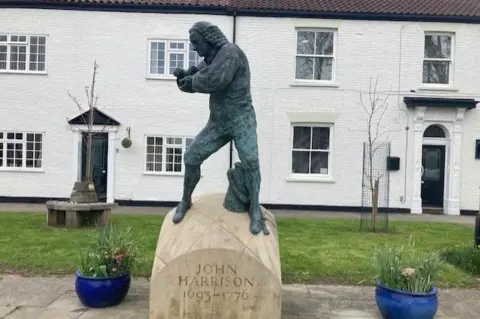 A dark grey statue of a man in 18th Century clothing and wig. He is looking down at an object held in his hands. A beige stone plinth has the words "John Harrison, 1693-1776" etched into it. The statue stands in front of a patch of green grass and a white-washed terrace of houses. Blue planters stand to either side of it.