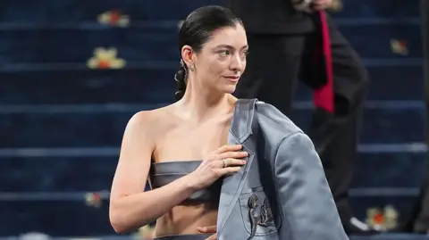 Getty Images Lorde at the Met Gala in New York. The singer wears a grey-blue silk blazer over a backless bandeau top and tight skirt. Her dark hair is pulled back in a plait and she wears a simple make-up.