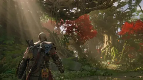 Sony A bald character clad in leather armour and with a large axe on his back walks into a forest scene. It's realistically illuminated with shafts of sunlight breaking through the canopy of the trees.