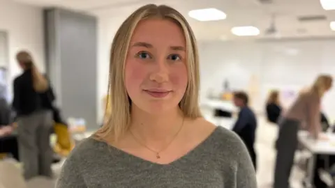 Elen has long, blonde hair and is wearing a grey jumper. She is standing in a large room where the new resources were launched, and behind her,  out of focus, are the people who attended the event talking to each other at tables.
