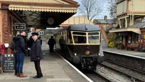 People mill about on an old-fashioned railway station on a cloudy day. Some men are wearing vintage guards uniforms. An historic Great Western Railway railcar painted in brown and white livery is pulling into the platform. 