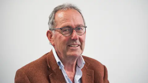 NHS Cornwall and Isles of Scilly Integrated Care Board A man, with short grey hair and glasses, smiling at the camera. He is wearing a blue shirt and brown blazer. Behind him is a grey background. 