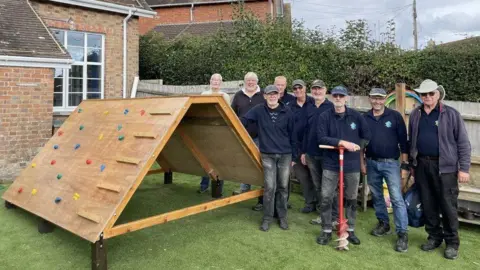A group of men are stod in a garden next to a children's climbing wall that has been constructed out of wood. The wall includes various coloured grips and a section of laddering. 