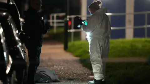 A forensic investigator in protective clothing examines an item on a pavement at night, illuminated by a light, with another person standing nearby.