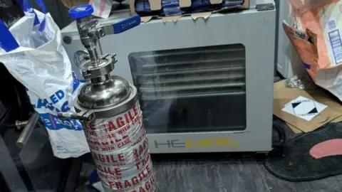 An old, grey oven and large silver gas cannister which is wrapped in tape with the words Fragile written in red capital letters. There are envelopes and plastic bags strewn around the scene. 