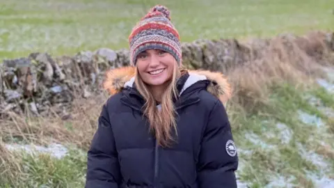 McInally family Hannah McInally, a young woman smiling at the camera, in a countryside setting, wearing a warm jacket and warm hat.