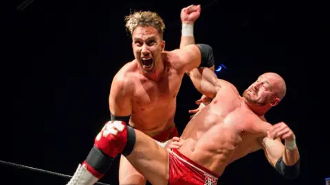 Emma Meek Two wrestlers, mid-match, mostly unclothed, wearing shorts and protection on knees and elbows. One looks to be screaming as it seems he is about to lift the other.