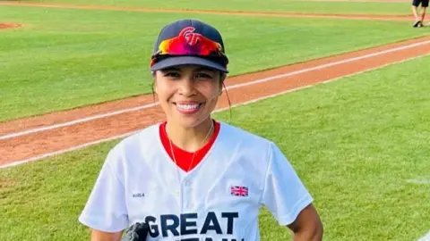 Betheliza Dela Paz stands on a baseball field with a Great Britain team shirt on and a blue cap. She has brown hair tied back and brown eyes and a big smile on her face.