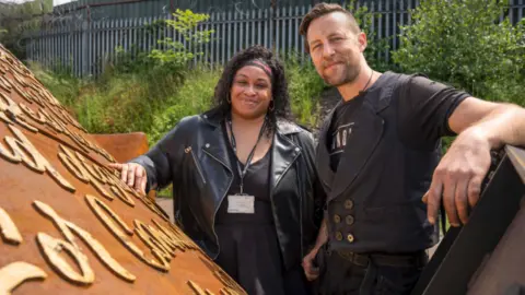 Victoria Murrain (left) and sculptor Luke Perry stand with the Windrush monument which is partially hidden from view in the park. What can be seen is a Corten steel colour with writing on it. The artists stand together behind it both wearing black and smiling at the camera. Victoria has long black hair and a scarf around her head. Luke has short brown hair and a beard.