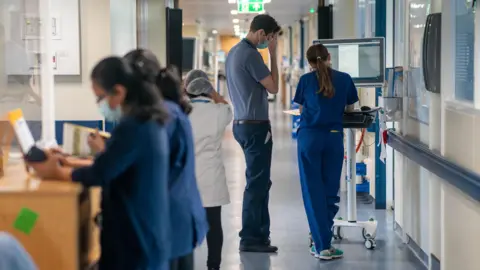 A group of five NHS workers engaged in tasks in a hospital corridor. Two women, dressed in blue uniforms, are examining documents while standing at a wooden desk. A woman wearing a white uniform has her back to the camera. A tall man dressed in a blue polo shirt and chinos stands by a computer station talking to a woman wearing a blue uniform.