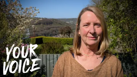 Grace Lloyd standing outdoors in Caerphilly county, in a sunny garden with trees and rolling hills in the background, wearing a brown top. The words “YOUR VOICE” appear on the right side of the image. Grace is shown from the waist up in a natural, outdoor setting.
