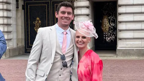 A man in a white suit with a pink tie is next to a woman dressed in pink with a pink fascinator on her head. They're both standing close to each other smiling.