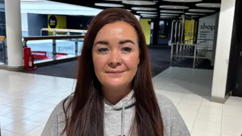 A woman with brown hair wearing a grey hoodie standing in an empty shopping centre.