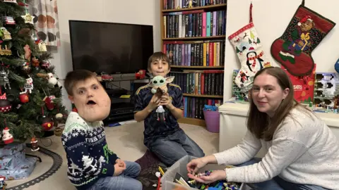 Alex playing in the living room with his mum Emily, and brother Roman. They are playing with Lego. Alex has a Minecraft themed Christmas jumper, and Roman is holding a toy of baby Yoda from Star Wars. A Christmas tree can be seen in the background, with a small model train running around its base.