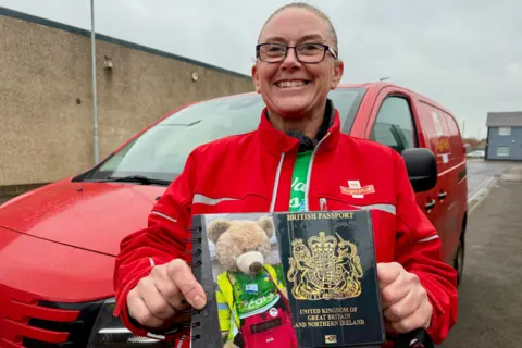 Nikki Gaynor has her blonde hair tied back and is wearing black-rimmed glasses and smiling. She's holding up a mock passport with a large teddy bear's face on the front. Behind her is a red post van.
