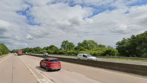 Google A motorway with a low concrete barrier in between the two carriageways. A red car is driving away from the shot and a white van towards it. The sky is blue in the background. 