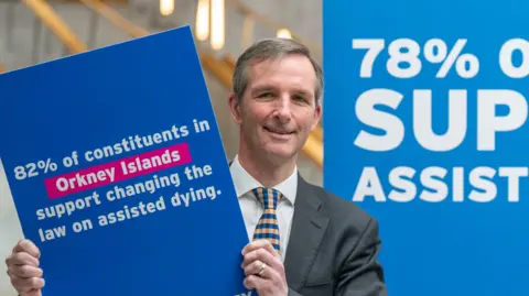 Liam McArthur - a man with brown hair and wearing a suit with a yellow and blue tie, holds up a placard saying people in the Orkney Islands support changing the law on assisted dying.