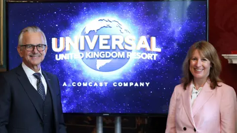 Toby Melville/PA Wire Mark Woodbury, on the left of frame, wearing a dark three piece suit and tie, with Rachel Reeves on the far right of frame wearing a buttoned up pink jacket and white blouse. Both are smiling broadly. They are separated by a large TV screen, on which is the name and logo of the Universal United Kingdom Resort. 
