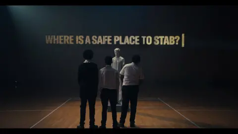 StreetDoctors/Saatchi&Saatchi In a still from the campaign film, three teenage boys stand with their backs to us in a sports hall, looking at a male mannequin, and a film screen which bears the words 'Where is a safe place to stab?' 