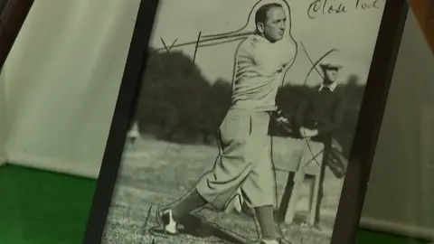 Old black and white photo of Bobby Cruickshank swinging a golf club, with trousers tucked into socks, and a smartly-dressed man looks on in the background