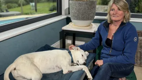 A woman sits wearing a blue top in a room with two windows and a large vase behind.