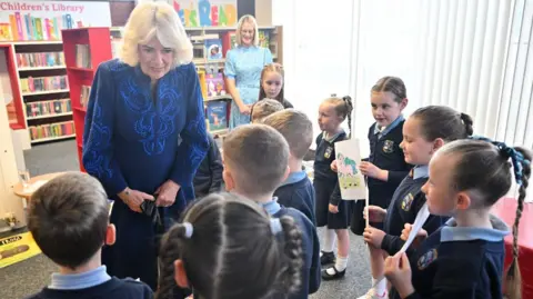 PA Media Queen Camilla is in a blue dress and is speaking with a number of school children. Some of the children, all dressed in uniforms, are holding pictures they have painted. A teacher in a blue dress and wearing glasses stands in the background. 