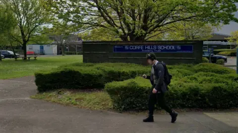 Google A school boy walks past the blue entrance sign to Corfe Hills School. The sign is mounted on a brick wall surrounded by shrubs.