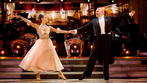 Two people, Wynne Evans and Katya Jones, stand on a stage. They are dancing. He is wearing a black suit and she is wearing a peach-coloured ballgown and heels.