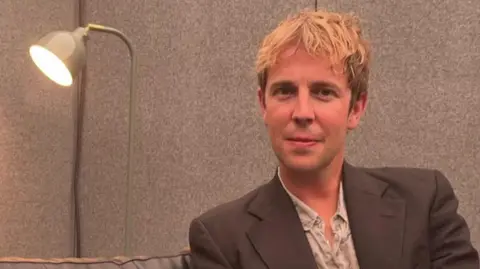 A head-and-shoulders image of Tom Odell sitting next to a lamp. He is smiling and looking into the camera.