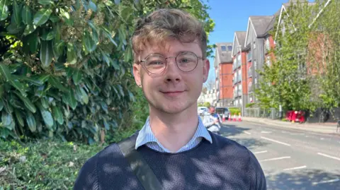 Kate Bradbrook/BBC A man with light hair and glasses is stood on a street