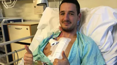 Rob Belt A man who is lying in a hospital bed giving a thumbs up. He has dark short hair and there is a large dressing on the right side on his chest.