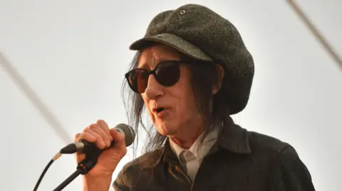 Jim Dyson/Getty Images A man holding a microphone, which is on a microphone stand. He is wearing glasses and a hat. 
