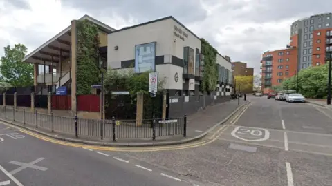 A road junction in London E14. There are modern red apartment blocks at the end of the road and a modern school building at the junction. The number 20 indicating the speed limit is painted on a speed bump in the road. 