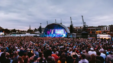Lloyd's Amphitheatre in Bristol. The stage is a semi circle shape and it is lit up with pink and blue spotlights. There is a large crowd of people standing and watching the performance. The Bristol landscape can be seen in the distance behind the stage.