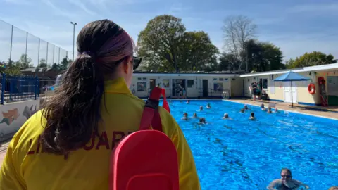 A lifeguard has her back to the camera as she watched over the pool. The water is clear and the sky is blue. There are about 20 swimmers in the water.