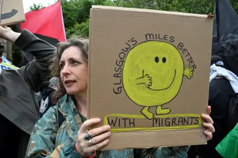 PA Media A woman holiding a cardboard sign featuring a yellow Mr Happy and the slogan: "Glasgow's miles better with migrants".