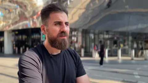 A bearded man with dark hair and a black t-shirt stood on a roadside in Birmingham city centre