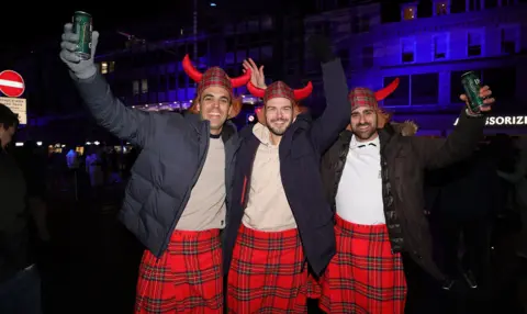 PA Media Three men in red kilts and tartan viking helmets holding cans of beer