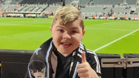 Family Handout Alfie Pentony, wearing black and white Newcastle United football top, with scarf round his neck, giving a thumbs up, in front of football pitch.