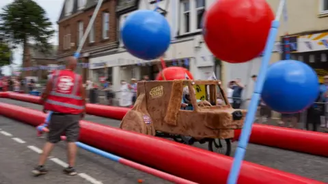 Stuart White A wooly brown car, centre, with ears and a nose resembling a dog rushing down a street. The background is blurred showing its speed. There is a bike wheel at the bottom. The car is dodging blue and red balls swinging from a frame above it.