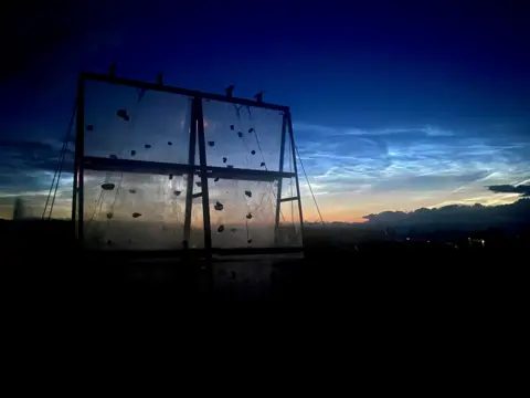 Richard Taylor A twilight landscape photograph featuring a climbing wall silhouetted against a luminous evening sky, with sky lit up by noctilucent clouds - high-altitude clouds that remain illuminated long after sunset.