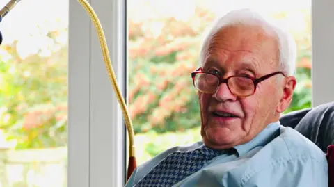 John Dockerty, a man who is sitting down in a chair with large glass windows behind him. He is sitting to the side of the picture and is looking directly at the camera. He is wearing a blue shirt with a blue dogstooth patterned tie and a pair of glasses.