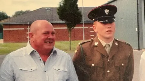 Joe Hemming Two men are stood next to each other in an old photograph. A man on the left is pictured wearing a light blue shirt with a bald head while a man on his left is pictured in a brown military uniform and black military cap.