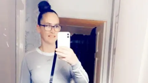 Other Kelly wearing a grey top and black glasses with her hair in a bun. She is holding a white phone as she smiles in a mirror.