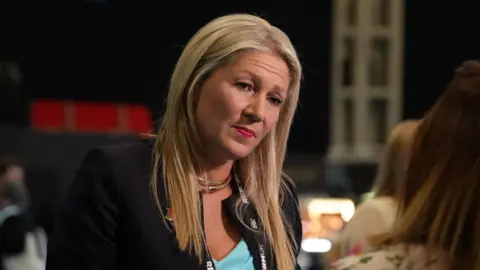 Sam Robinson/BBC Laura Anne Jones speaking to an individual off camera, wearing a black jacket and blue top.