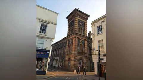 Victorian Society The ornate brown, black and orange brickwork building stands between other newer buildings in the town. Two cream buildings stand either side with pedestrians walking below. The building is a large square shape with a tall tower in the centre.