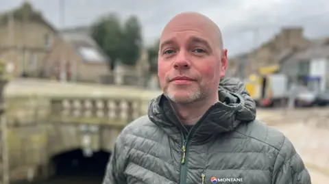 A bald-headed man with short grey beard, wearing a green jacket, looks at the camera with a street in the background