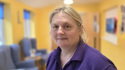 Paul Moseley/BBC Melanie Baldwin is wearing a purple polo-neck shirt. She is standing in a corridor with yellow walls.