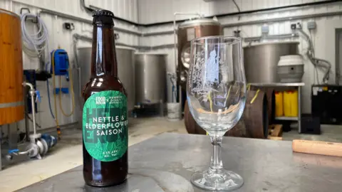 Ben Schofield/BBC A bottle of Humber Doucy beer and an empty half pint glass, which also has the brewery's name and logo on it. Both are sitting on a metal table in the brewery's warehouse. Brewing machinery can be seen in the background.